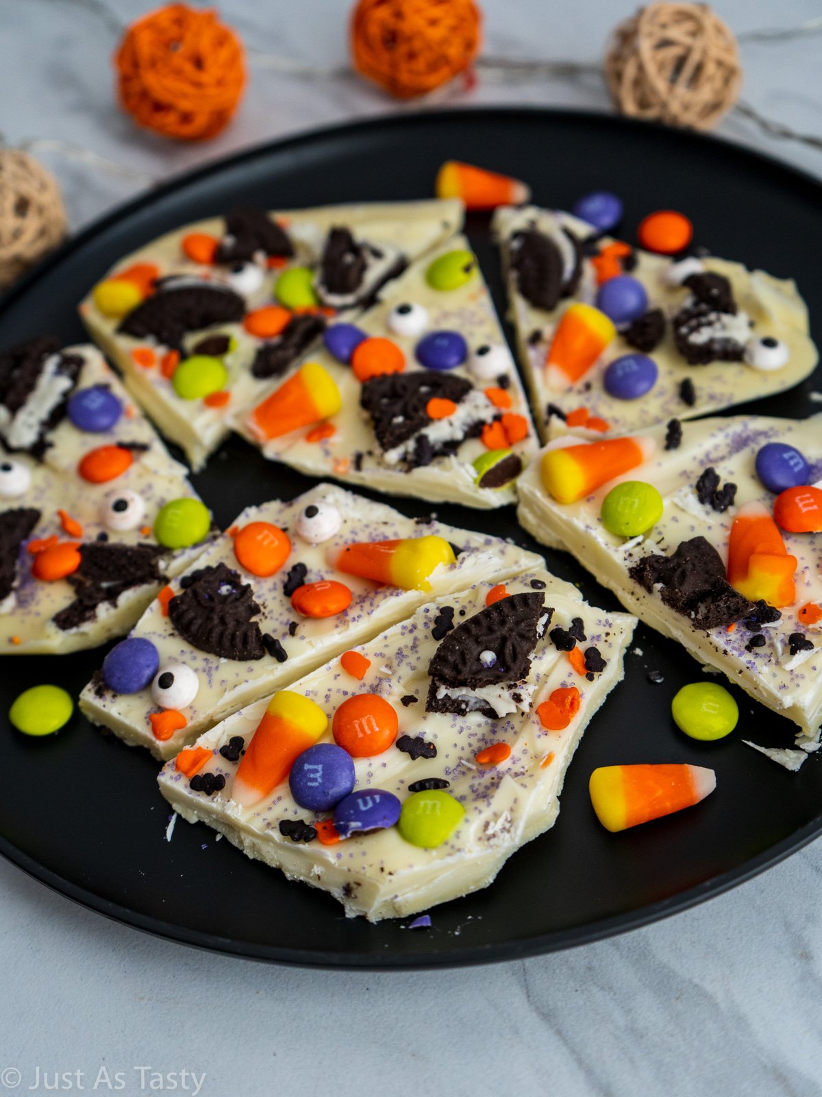 White chocolate bark topped with candy corn, Halloween sprinkles, and Oreo pieces on a black plate. 