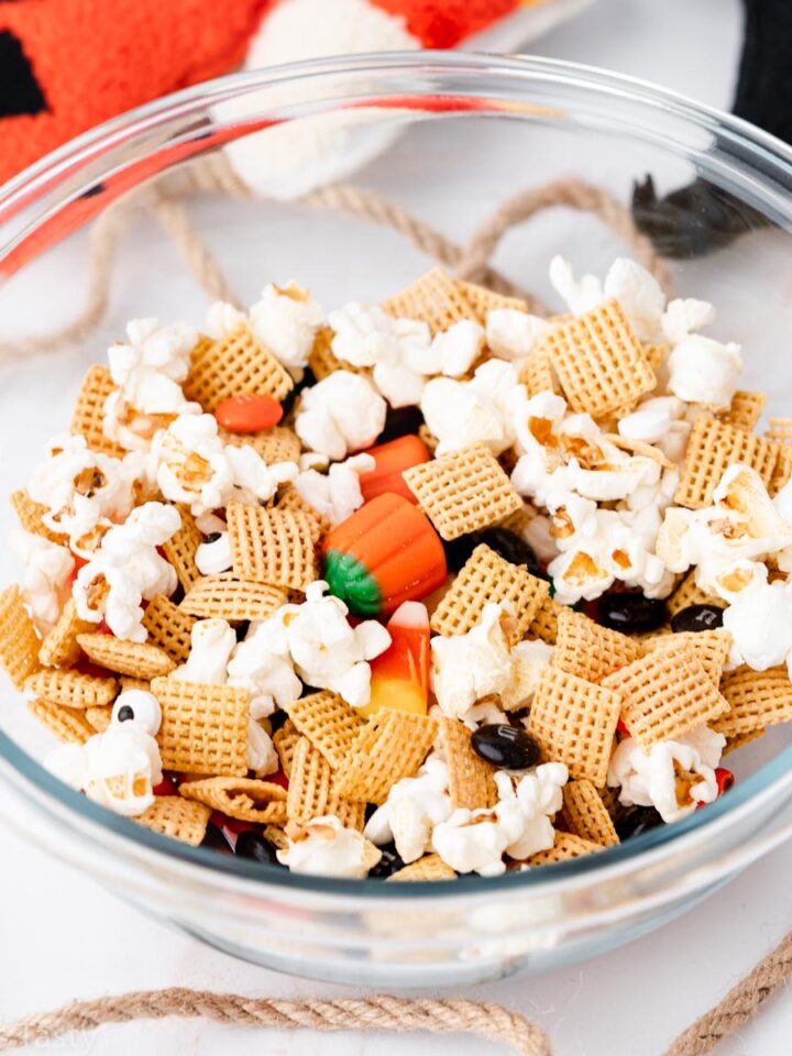 Halloween snack mix of candy corn, popcorn, Corn Chex cereal, and M&Ms in a glass bowl.
